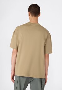 Joven con cabello corto que lleva una camiseta de manga corta beige y suelta, y pantalones grises, de espaldas frente a un fondo blanco liso.