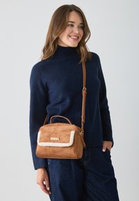 Sac à main en daim tan avec un rabat en fourrure crème, fermeture éclair et une bandoulière en cuir amovible. Le sac possède une petite poignée sur le dessus.