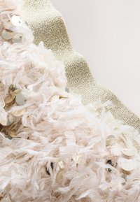 Svjetloružičasti teksturirani materijal s poderanim rubovima, raspršenim zlatnim šljokicama i akcentom metalik zlatne vrpce duž gornjeg ruba.