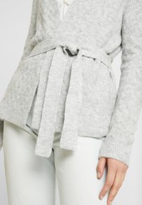 Ljusgrå, kabelstickad cardigan med knytband. Ribbad mudd. Bärs över ljusgröna byxor. Mjuk, texturerad tyg.