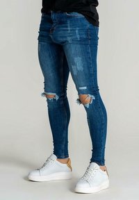 Úzké modré džíny z denimu s opranými a roztrhanými koleny, s přiléhavým střihem. Nošeny s bílými teniskami s hnědými detaily.