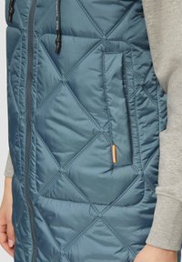 Ge quiltete Weste in Teal, mit durchgehendem Reißverschluss vorne und Seitentasche. Das Material hat eine glatte Textur mit rautenförmigen Steppnähten.