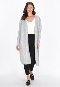 Femeie zâmbitoare îmbrăcată cu un cardigan lung, gri deschis, cu mărgeluțe, o bluză albă cu decolteu în V, pantaloni negri și mocasini negri, mergând înainte pe un fundal alb.