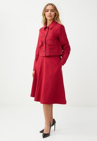 Veste rouge avec deux poches poitrine et une jupe évasée assortie, fabriquée en tissu lisse, accompagnée de chaussures à talons hauts noires.