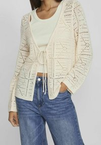 Cárdigan de punto abierto color crema con patrones geométricos, un cierre de lazo en la cintura y mangas largas, combinado con una camiseta sin mangas clara y jeans azules.