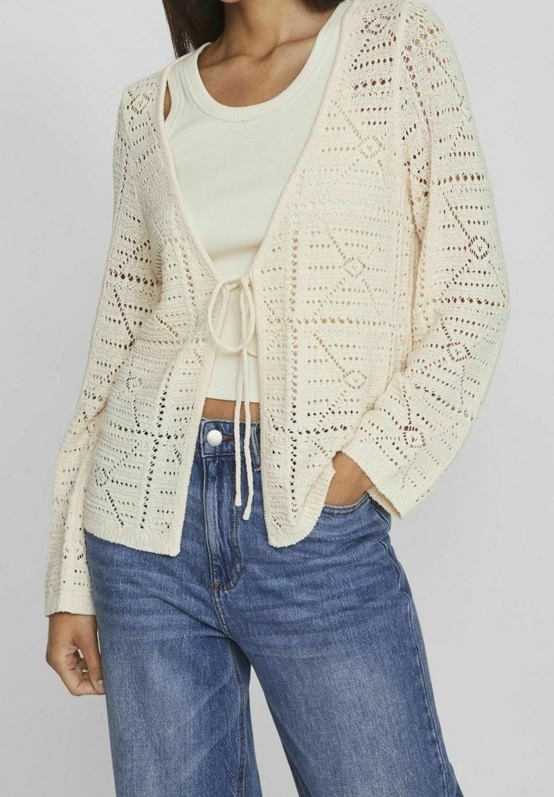 Cárdigan de punto abierto color crema con patrones geométricos, un cierre de lazo en la cintura y mangas largas, combinado con una camiseta sin mangas clara y jeans azules.