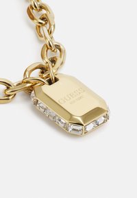Guld kæde med en sekssidet vedhæng, der har et indgraveret mærkenavn og en klar krystal-accentueret kant, glat overfladetextur.