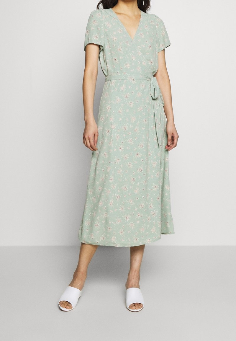 Femme portant une robe midi portefeuille fleurie vert clair à manches courtes et des sandales blanches ouvertes à l'avant, debout devant un fond uni.