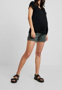 Svart kortärmad topp med v-ringning och subtil spetsdetalj, kombinerad med gröna uppvikta shorts och svarta sandaler med remmar.