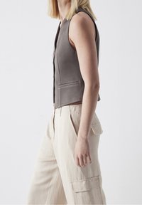 Szara bezrękawnik z wysokim dekoltem, z przodu zapinany na guziki i z bocznymi kieszeniami, w połączeniu z jasnobeżowymi, szerokimi spodniami.