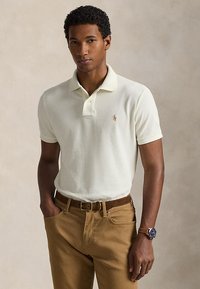 Mann, der ein weißes Polohemd, hellbraune Hose mit braunem Gürtel und eine Armbanduhr trägt, steht mit einer Hand in der Tasche vor einem einfarbigen Hintergrund.