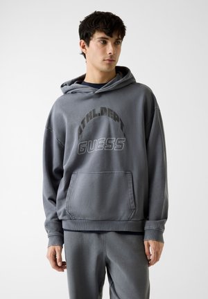 Felpa grigia con cappuccio, tasca frontale, vestibilità comoda e testo in grassetto "ATHL.DPT" e "GUESS" in colori a contrasto.