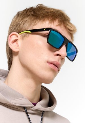 Giovane con capelli corti castano chiaro che indossa grandi occhiali da sole neri con lenti riflettenti blu e una felpa beige con cappuccio.