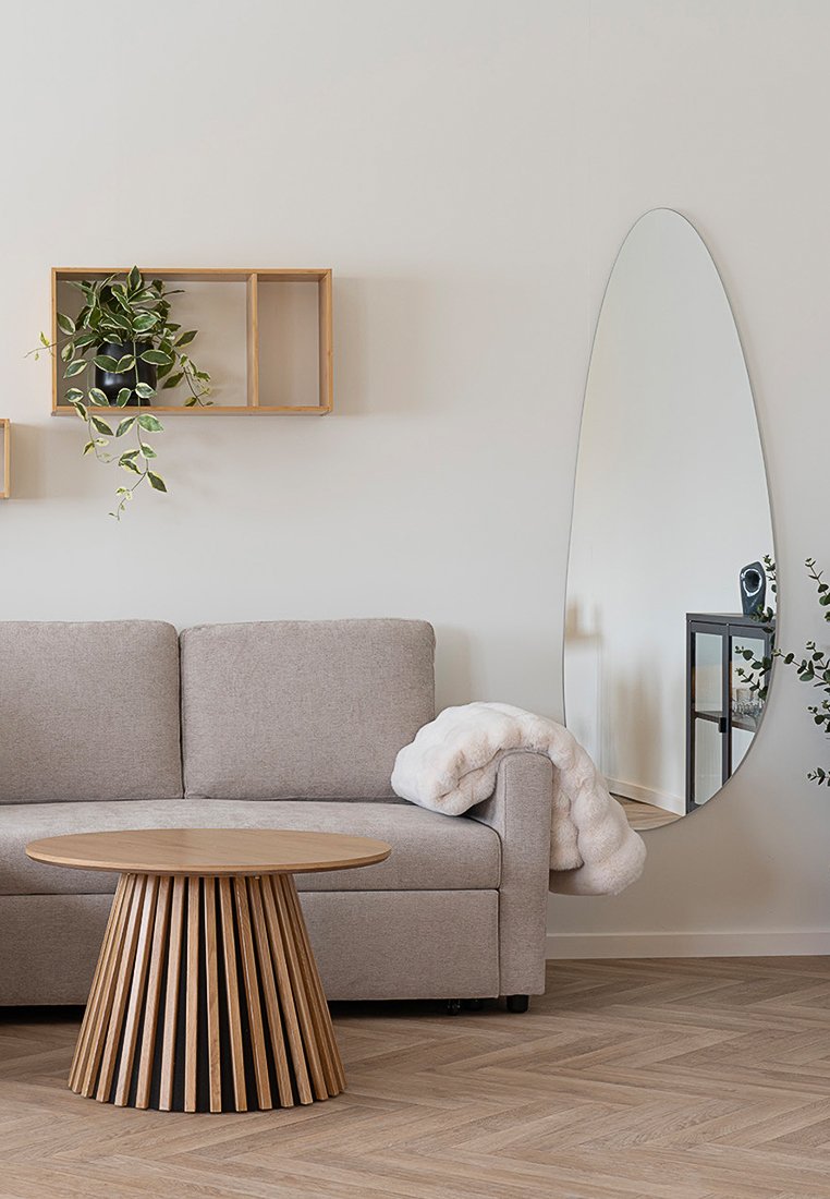 Specchio ovale con una cornice sottile, divano in tessuto grigio, tavolo rotondo in legno con doghe verticali e mensola per piante a muro. Morbido plaid beige appoggiato sul divano.