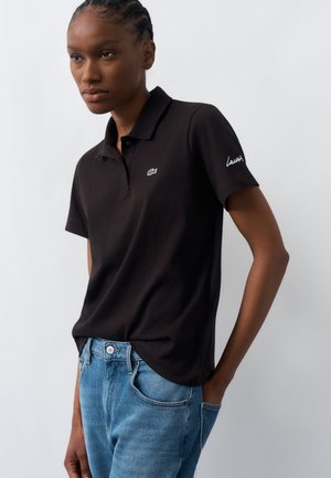 Femme portant un polo noir avec un petit logo vert sur la poitrine et un jean bleu, debout avec une main dans la poche devant un fond uni.