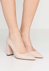 Escarpins en cuir verni beige à talon aiguille, avec un bout pointu, un talon carré et une texture lisse. Mettent en valeur un design épuré et un détail minimal.