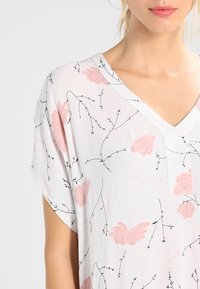 Haut à col en V et manches courtes en tissu blanc léger. Présente un motif floral rose et des accents de lignes noires. Texture douce.