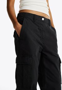 Pantalones cargo negros de corte relajado, con bolsillos laterales, cierre de botón y una tela suave y texturizada. Costuras visibles como acentos.