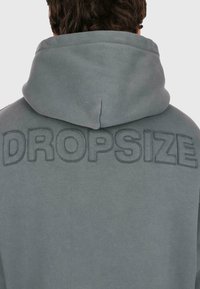 Mann trägt ein graues Kapuzenpullover mit erhabenem, gesticktem "DROPSIZE"-Text über dem oberen Rücken.