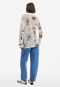 Baltas, raukšlėtas kardiganas su spalvingais gėlių ir raštų motyvais. Dėvimas su mėlynomis laisvo kirpimo džinsomis ir šviesiais, spalvotais batais.