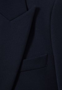 Detail tmavomodrého materiálu saka ukazujúci golier a pásik vrecka.