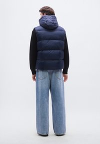 Colete acolchoado azul-marinho com capuz, apresentando secções acolchoadas. Usado sobre um suéter preto e combinado com jeans azul claro de perna larga.