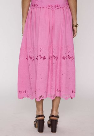 Femme portant une jupe midi rose avec broderies florales et détails découpés, associée à des sandales à talons hauts marron foncé et des bracelets en or.