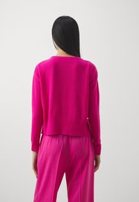 Frau mit langen schwarzen Haaren, die einen leuchtend pinken Pullover und passende pinke Hosen trägt, von hinten vor einem schlichten hellen Hintergrund gezeigt.