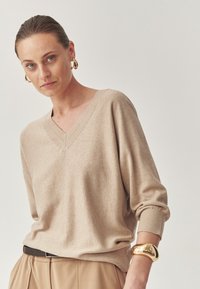 Beige V-nyakú pulóver puha kötésből, bordázott mandzsettákkal, bézs nadrággal és fényes arany csuklószorítóval párosítva. Egyszerű, laza szabású.