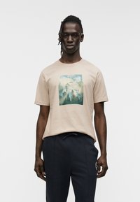 Ανοιχτό μπεζ βαμβακερό μπλουζάκι με γραφικό σχέδιο σε πράσινο και μπλε στο μπροστινό μέρος. Κοντά μανίκια, στρογγυλή λαιμόκοψη, σε συνδυασμό με σκούρο ναυτικό παντελόνι.