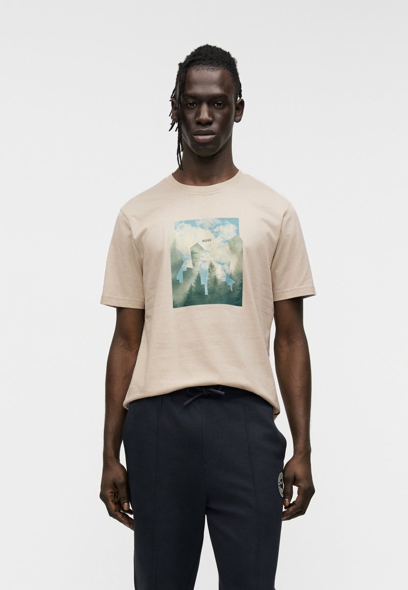 Ανοιχτό μπεζ βαμβακερό μπλουζάκι με γραφικό σχέδιο σε πράσινο και μπλε στο μπροστινό μέρος. Κοντά μανίκια, στρογγυλή λαιμόκοψη, σε συνδυασμό με σκούρο ναυτικό παντελόνι.