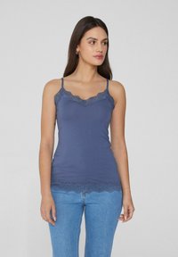 Ciemnoniebieski top na ramiączkach z koronkowymi detalami przy dekolcie i u dołu, z cienkimi, regulowanymi ramiączkami. Noszony z jasnoniebieskimi dżinsami.