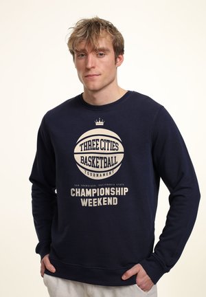 Felpa navy con maniche lunghe, caratterizzata da una grafica di pallacanestro color crema e dal testo: "Torneo di Pallacanestro delle Tre Città - Weekend del Campionato."