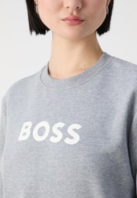 Personne aux cheveux courts et foncés portant un sweat-shirt gris avec un texte blanc BOSS, montré du menton jusqu'à la partie supérieure de la poitrine sur un fond uni.