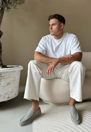 Jonge man in een witte outfit en grijze instappers zittend op een gestructureerde stoel naast een grote potplant tegen een beige muur.