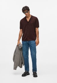 Ruda trumpa rankovėmis marškinėliai su mygtukais ir siuvinėjimu, derinami su mėlynomis džinsomis ir juodais batais, laikant tekstūruotą pilką švarką.