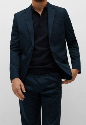 Hombre vestido con un traje azul marino oscuro, con americana y pantalones a juego, combinado con una camiseta polo negra, posando frente a un fondo liso.