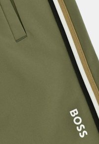 Pantalons de jogging vert olive avec une texture lisse, dotés d'une poche zippée sur le côté avec des stripes noirs, blancs et dorés, et un logo "BOSS" blanc.