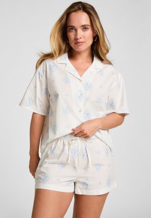 Jeune femme aux cheveux blonds portant un haut crème à manches courtes boutonné et un short assorti avec un imprimé floral bleu clair sur fond gris.