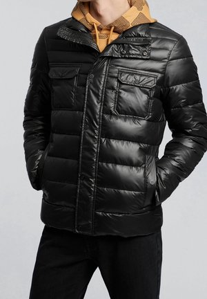 Homme portant une veste matelassée noire sur un sweat à capuche moutarde avec motif géométrique, mains dans les poches de la veste, sur fond clair.