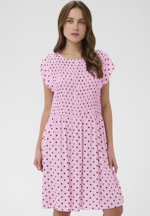 Młoda kobieta ubrana w różową sukienkę do kolan w brązowe kropki z krótkimi rękawami, stojąca na jasnym, jednolitym tle.