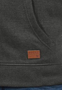 Mørkegrå hættetrøje med en tekstureret overflade, der har et brunt lædermærke med påskriften "BLEND GLOBAL JEANMAKER" nær kanten.