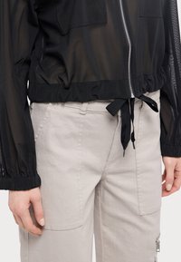 Chaqueta negra semi-translúcida con cierre de cremallera y cintura elástica, combinada con pantalones grises claros que cuentan con bolsillos y perneras rectas.