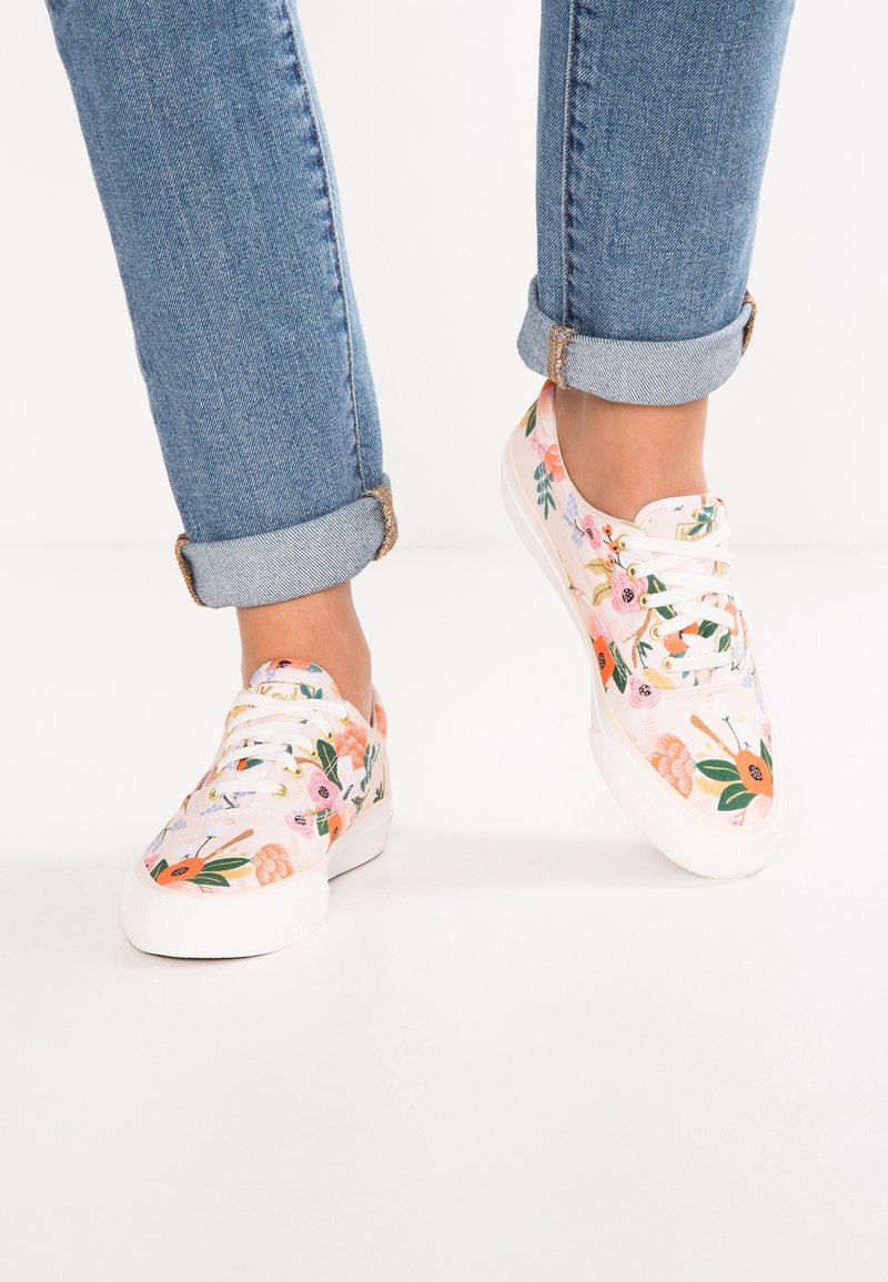 Personne portant un jean bleu clair retroussé et des baskets blanches avec des motifs floraux colorés sur un fond blanc.