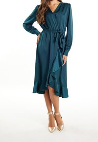 Femme portant une robe longue à manches longues de couleur teal avec une encolure croisée et un ourlet volanté, se tenant avec une main sur la hanche sur fond blanc.