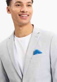 Lichtgrijze blazer van een textuurstof, met een inkeping op de revers en een felblauwe pochet. Draagover een wit T-shirt.