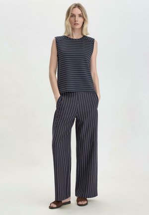 Top senza maniche blu navy con righe bianche, abbinato a pantaloni larghi blu navy con sottili strisce verticali. Infradito marroni.