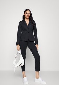 Zwarte op maat gemaakte blazer met twee knopen, gecombineerd met slim-fit zwarte broeken. Een witte schoudertas en witte sneakers maken de outfit compleet.