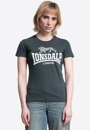 Žena s krátkymi blond vlasmi, ktorá má na sebe tmavozelené tričko Lonsdale London a modré rifle na jednoduchom bielom pozadí.