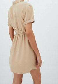 Beżowa sukienka z krótkim rękawem, z kołnierzykiem, zebranym w talii i lekko rozkloszowanym dołem, wykonana z lekkiego, gładkiego materiału.
